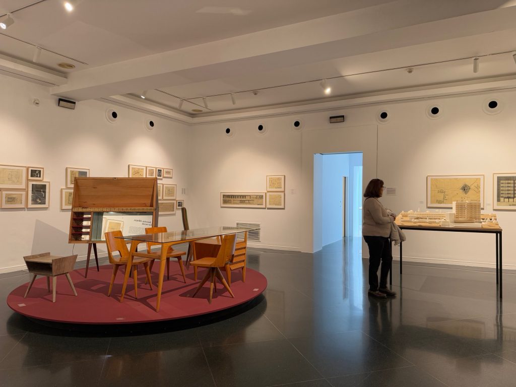 Sala de la exposición con mobiliario sobre una tarima circular y planos en las paredes