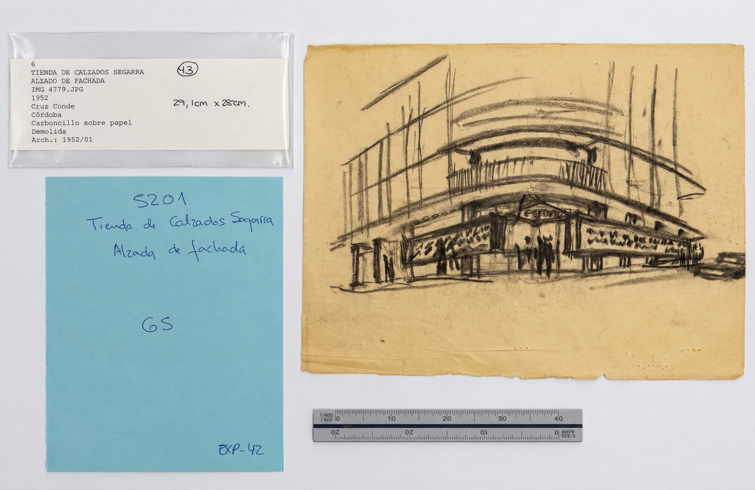 Dibujo a carboncillo de la Tienda de Calzados Segarra junto a su etiqueta de catalogación, una nota manuscrita y una regla, sobre la mesa de trabajo del archivo